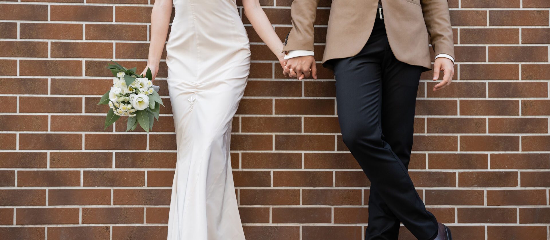 cropped view of young bride in white dress holding wedding bouquet and hand of groom near brick wall